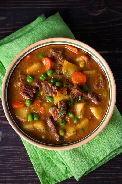 Homemade Irish beef stew with potatoes, carrots and peas in bowl on wooden table. overhead, vertical