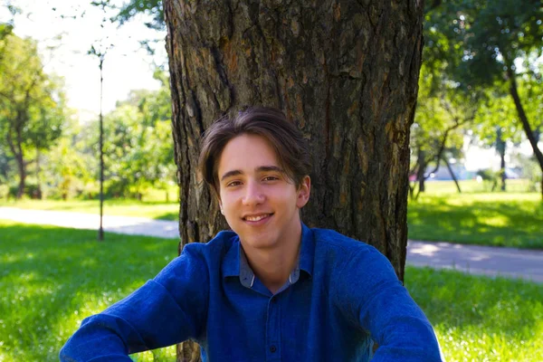 公園で木の横に芝生に座っている若い男 カメラを見て — ストック写真