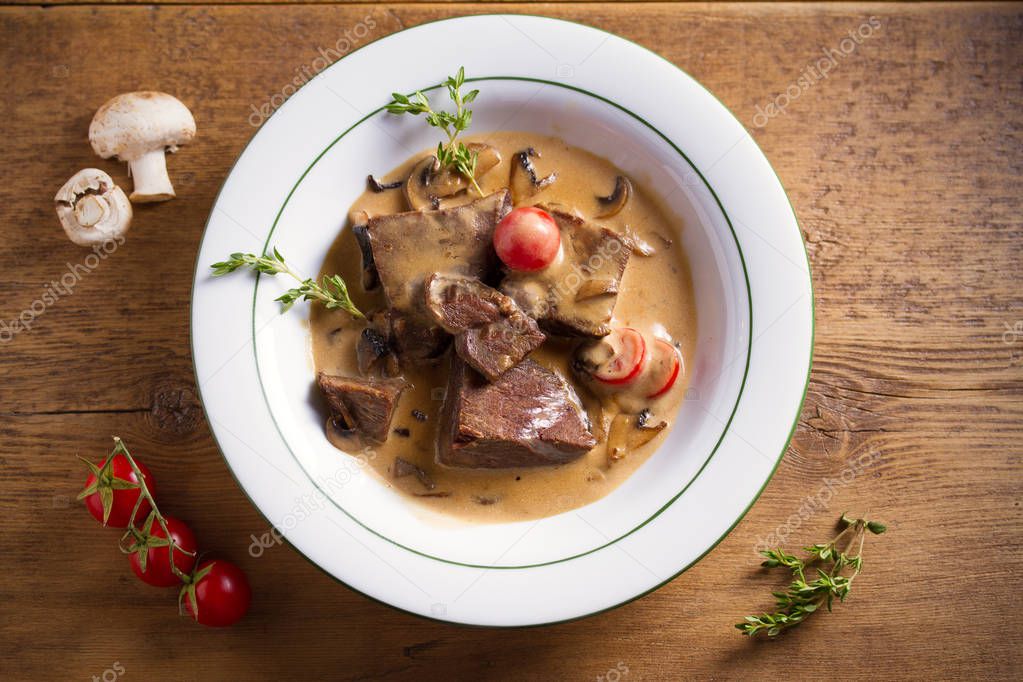 Braised beef cheeks in cream sauce with mushrooms and tomatoes in white bowl on wooden table. Overhead