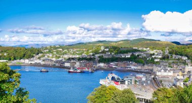 A panoramic view of Oban on the west coast of Scotland, showing the town, ferry terminals and hills in  the background, taken on a sunny day in early autumn.