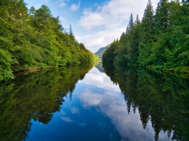 Laggan Avenue on the Caledonian Canal in Scotland, UK - on a calm, sunny day the sky and trees are reflected in the calm water of the canal. The Great Glen Way runs through the trees on the left. clipart
