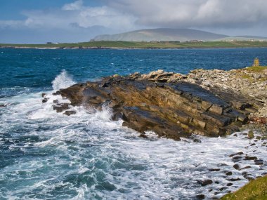 Sumburgh Head yakınlarındaki güney Shetland sahilindeki eğimli kumtaşı kayalarında dalga hareketi sağdaki dalgaların kaya kalıntılarını süpürdüğünü gösteriyor. Arka planda Fitful Head tepesi var..