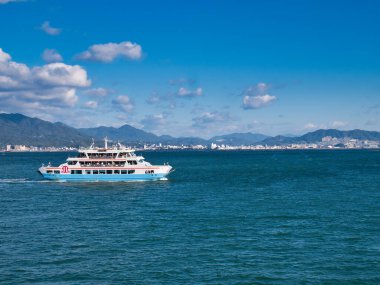 Miyajimaguchi İstasyonu ile Hiroşima Körfezi 'ndeki Miyajima Adası arasında yolculuk yapan yolcu dolu bir vapur. Arka planda Hiroşima 'nın banliyöleri görünüyor..