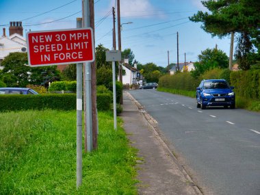 Kırmızı bir işaret, sürücüleri İngiltere 'nin Lancashire kentindeki Halsall kırsalında hız sınırının 30mph olduğu konusunda uyarıyor. Sağdan mavi bir araba geçiyor..