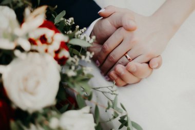 Gelin ve damat düğün töreninde düğün buketinin önünde el ele tutuşuyorlar. Koca, karısının elini nazikçe sıkar.
