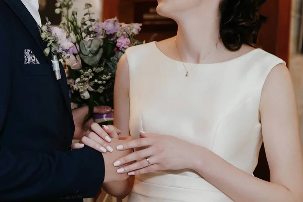 sweethearts hold hands standing at the wedding ceremony with a bouquet of purple flowers. a fist hangs on the bride, and a buttonhole on the bride