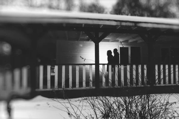 A silhouette of a hugging couple standing in the evening on a wooden veranda during a winter snowfall. black and white photography