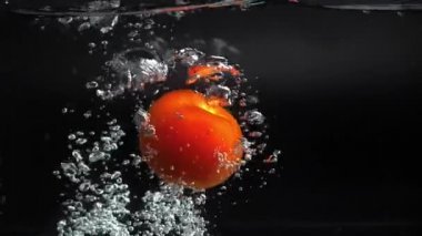 Büyük kırmızı bir domates suya su sıçratarak düşüyor, yavaşça dibe batıyor, arkasında bir sürü hava kabarcığı bırakıyor. Stüdyo çekimi siyah arka planda. Hızlı 180 fps