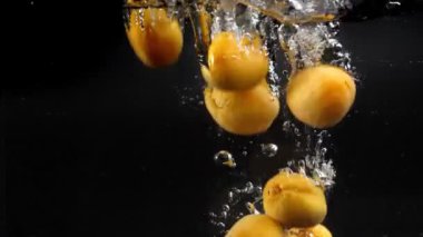 Birçok sarı kayısı suya spreyle düşer, yavaşça dibe batar, arkasında bir sürü hava kabarcığı bırakır. Stüdyo çekimi siyah arka planda. Hızlı 180 fps