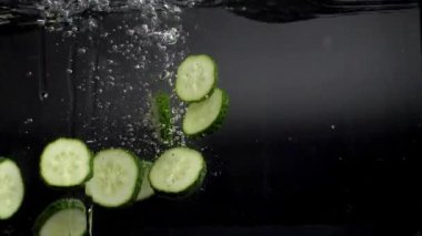 Bir sürü yeşil salatalık suya spreyle düşer, yavaşça dibe batar, arkasında bir sürü hava kabarcığı bırakır. Stüdyo çekimi siyah arka planda. Hızlı 180 fps