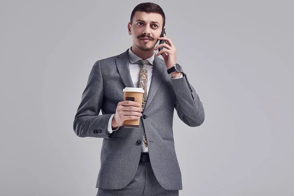 Portrait of handsome young confident arabic businessman with fancy mustache in fashion gray full suit holds a cup of coffee and talking on the phone