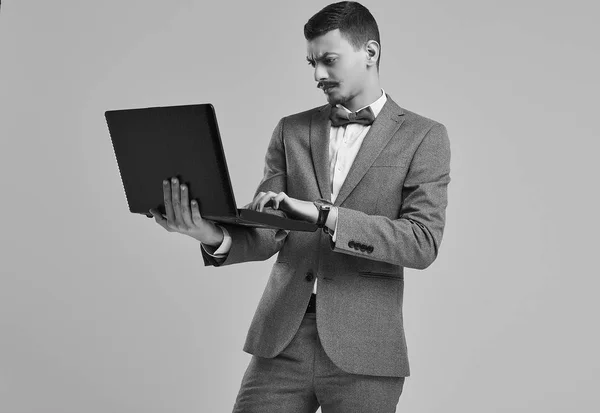 英俊的年轻自信的阿拉伯商人的肖像与花哨的胡子在时尚灰色全西装举行黑色笔记本电脑在工作室的背景 — 图库照片