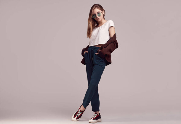 Портрет милой девочки-подростка в модной куртке и цветных очках, позирующих на сером фоне. Студийные съемки
.