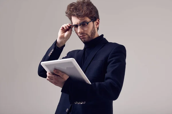 Fashion portrait of handsome elegant man with curly hair wearing suit and glasses holding a tablet on gray background in studio