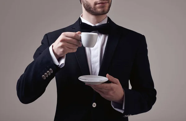 穿着燕尾服的帅气优雅男人的时尚肖像 穿着燕尾服 在工作室里拿着一杯灰色背景的浓咖啡 — 图库照片