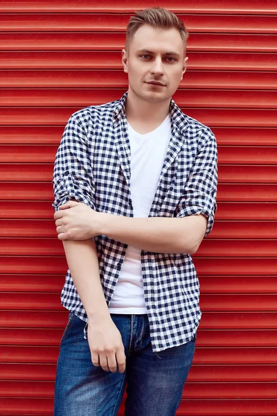 Joven hombre guapo en una camisa y jeans cerca de la puerta roja —  Fotos de Stock