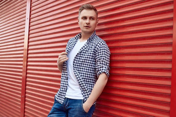 Jovem homem bonito em uma camisa e jeans perto do portão vermelho — Fotografia de Stock