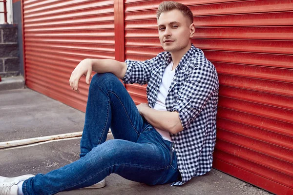 Joven hombre guapo en una camisa y jeans cerca de la puerta roja —  Fotos de Stock