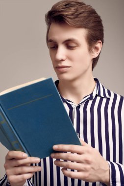 Çizgili gömlekli genç adam gri arka plan üzerine bir kitap okuyor.