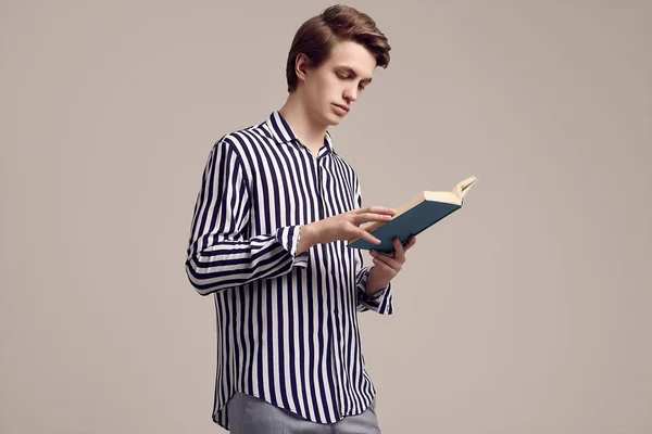 Jonge knappe man in gestreept shirt het lezen van een boek over grijze achtergrond — Stockfoto
