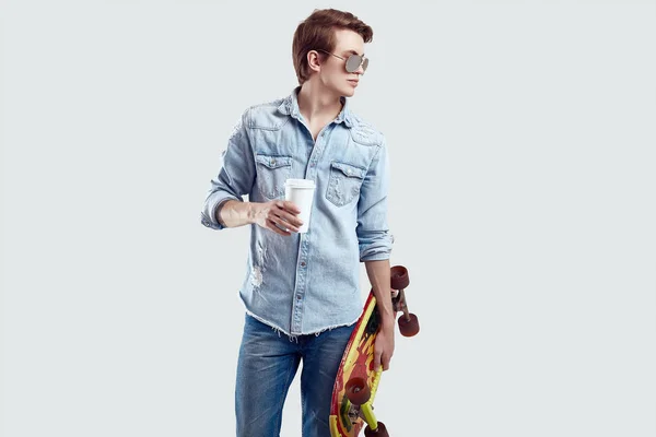Hipster hombre en gafas de sol y jeans chaqueta posando con monopatín y café —  Fotos de Stock