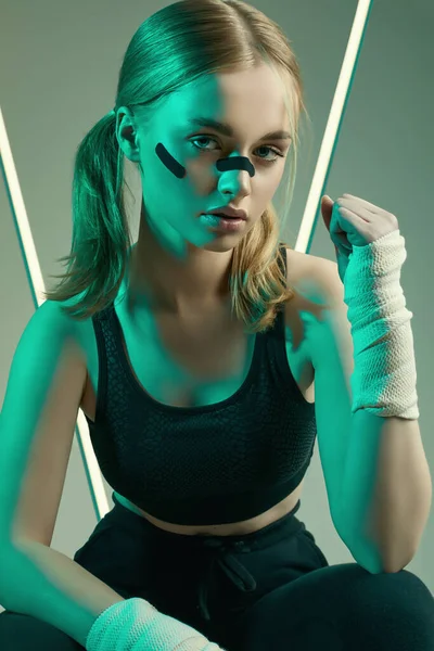 Vivid portrait of strong beautiful girl with blonde hair, sports figure, confident look and fists in protective boxing bandages posing on colorful neon light background