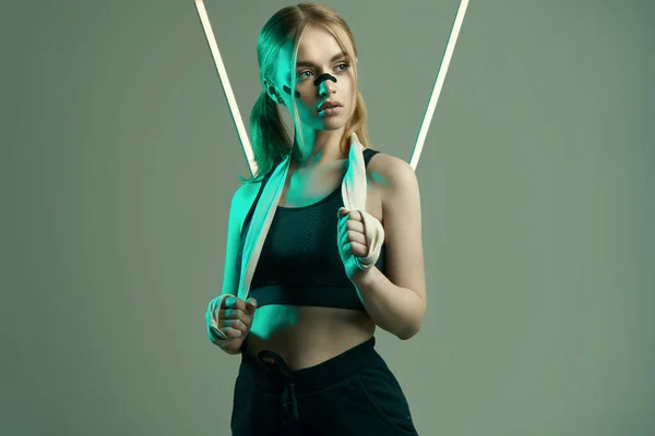 Vivid portrait of strong beautiful girl with blonde hair, sports figure, confident look and fists in protective boxing bandages posing on colorful neon light background