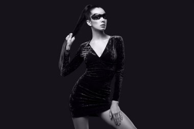 Güzel mor elbiseli ve pullu maskeli zarif esmer kadının portresi stüdyoda siyah arka planda poz veriyor.