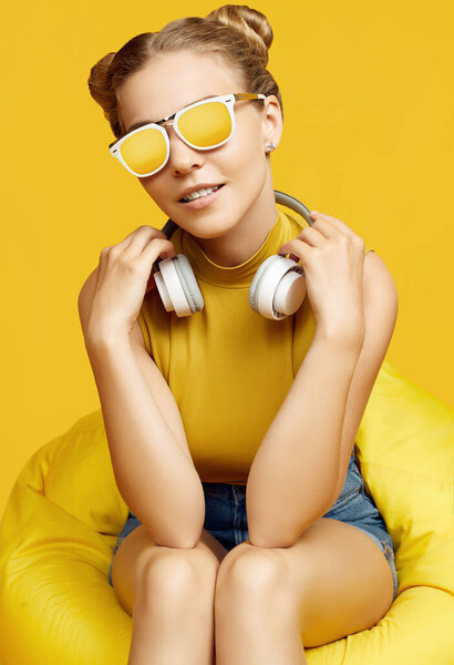 Portrait of gorgeous blonde hipster girl wearing sunglasses sitting in a sack bag with headphones on colorful yellow background