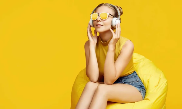 Portrait of gorgeous blonde hipster girl wearing sunglasses sitting in a sack bag with headphones on colorful yellow background