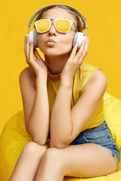 Portrait of gorgeous blonde hipster girl wearing sunglasses sitting in a sack bag with headphones on colorful yellow background