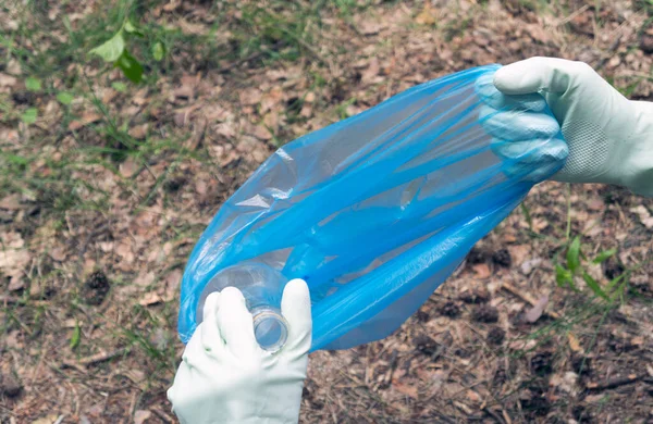 Man removes trash in nature. Hands in gloves put a plastic bottle in a trash bag. First-person view.