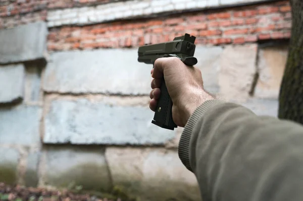 A gun in the hand of a Caucasian man against a brick wall of an abandoned building. Shooter with a gun of dark green color. First person view, pov.