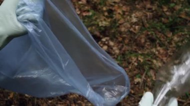 Eldiven giymiş genç bir adamın elleri plastik bir şişeyi çöp torbasına koyar. Ormanda çöp toplama.