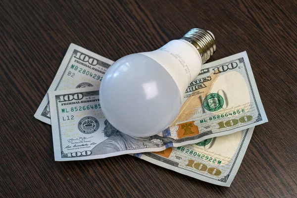 A light bulb and hundred-dollar bills lies on a brown wooden table. Electricity payment concept. Close-up.