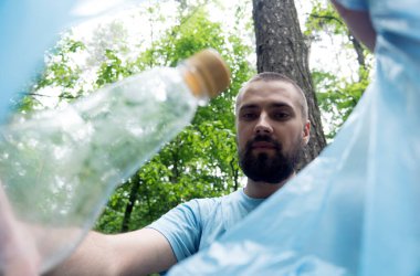 Sakallı genç beyaz bir adam çöp torbasına plastik bir şişe koydu. Poşetteki manzara. Doğada çöp toplama.