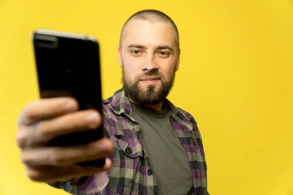 Portrait of bald and bearded caucasian man on yellow background. Online business concept with a copy space. Young guy holding phone in his hans in home office.