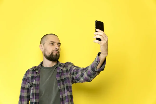 A young man with a beard and a bald head holding a phone in his hand on a yellow background. Blogger makes a selfie