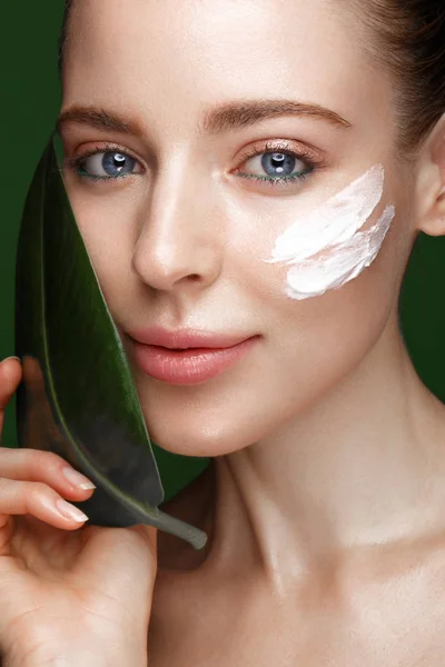 Beautiful fresh girl with cosmetic cream on the face, natural make-up and green leaves. Beauty face. — Stock Photo, Image