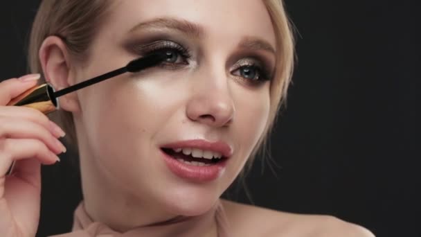 Beautiful blond girl with classic make up paints eyelashes with mascara, posing in the studio. Beauty face. — Stock Video