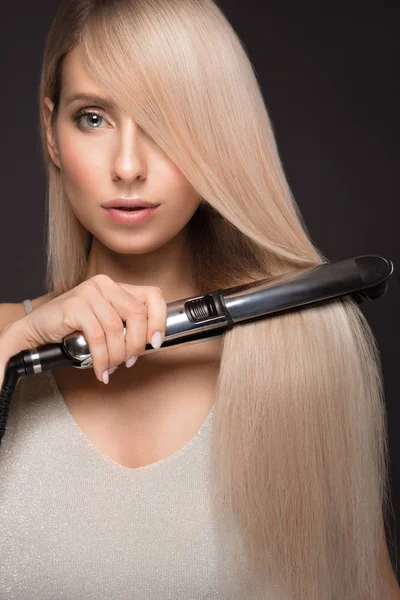 Belle fille blonde aux cheveux parfaitement lisses, maquillage classique avec un style de curling dans les mains . — Photo
