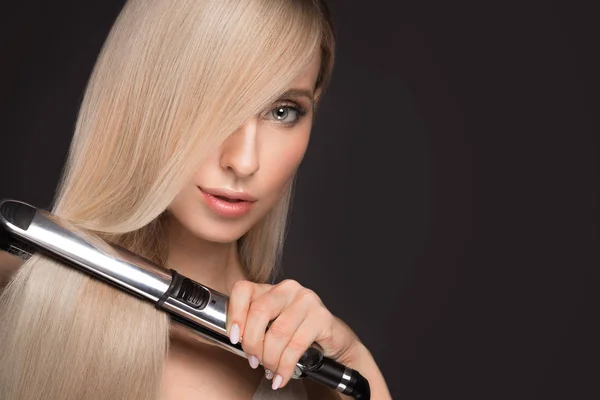 Belle fille blonde aux cheveux parfaitement lisses, maquillage classique avec un style de curling dans les mains . — Photo