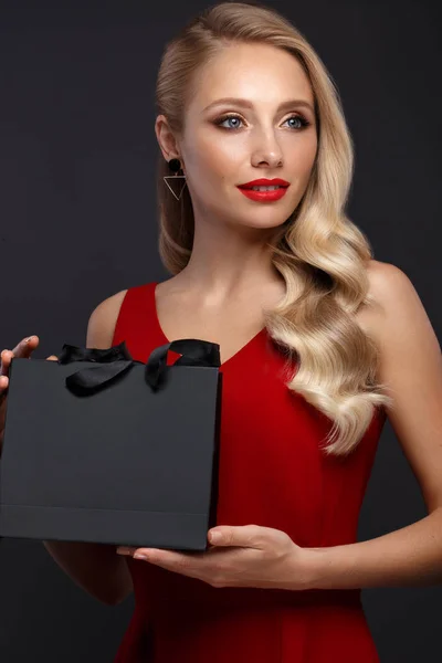Hermosa chica con cabello rubio, maquillaje clásico en un elegante vestido rojo y bolsa de compras. Cara de belleza . —  Fotos de Stock