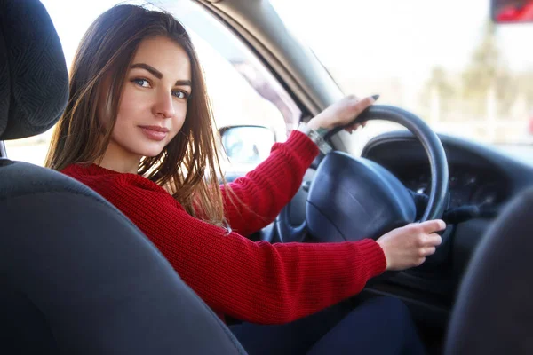Νεαρή όμορφη ευτυχισμένη γυναίκα στο κόκκινο κάθεται στο τιμόνι νέο εκπαιδευτικό αυτοκίνητο. — Φωτογραφία Αρχείου