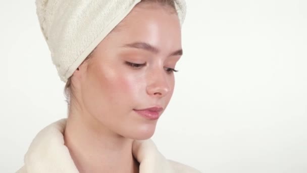 Piękna, delikatna młoda dziewczyna w białym ręczniku i szlafroku z czystą, świeżą skórą nakłada krem na twarz i pozuje przed kamerą. Piękna twarz. Skóra. — Wideo stockowe