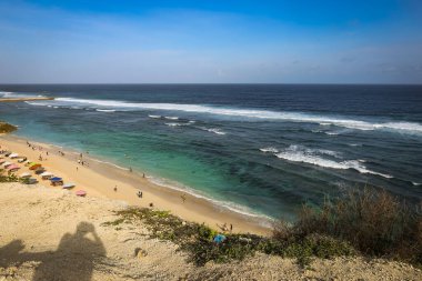 Pandawa Sahili 'nde geniş bir açı, Bali' de kumlu sahilde şemsiyeler, okyanusta güçlü dalgalar ve mavi bir gökyüzü.