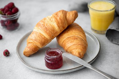 Gri bir masada ahududu reçelli taze kruvasan ve portakal suyu.
