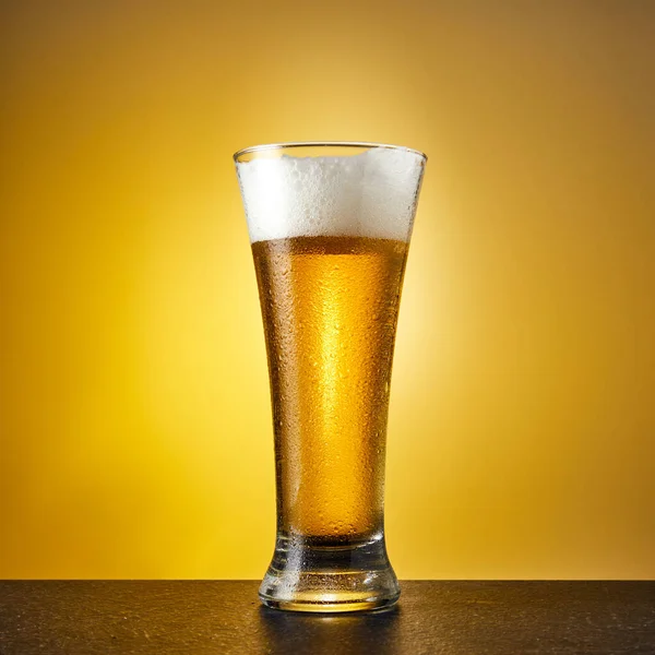 Karanlık bir masanın üzerine damlayan bardakta soğuk el yapımı hafif bira. Sarı arka planda yarım litre bira. Kapat..