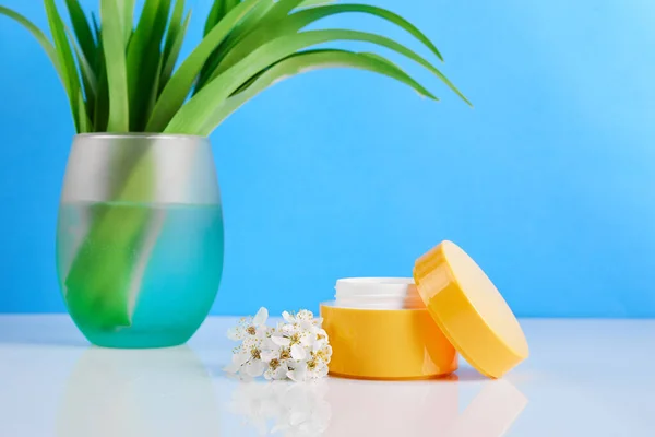 Jar of cosmetic cream and soft green vase with flowers on a blue background. Natural cosmetic product with herbs.