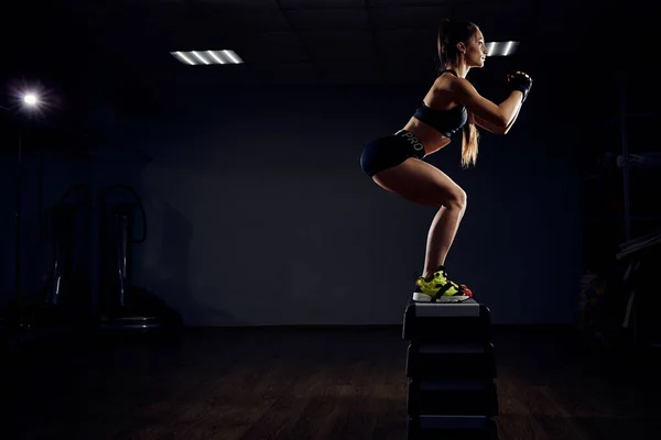 Zıplama egzersizi yapan zinde bir genç kadın. Kaslı kadın karanlık arka planda spor salonunda çömelme hareketi yapıyor. — Stok fotoğraf